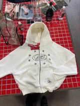 Buzo con cierre frontal, color crema, con diseño estampado de estrellas y logo en el pecho. Cuenta con capucha, bolsillos tipo canguro y tirador de cierre en forma de estrella.