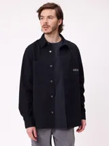 Camisa de gabardina color verde oliva, con cuello clásico, cierre frontal de botones y dos bolsillos frontales con parche. Incluye un pequeño parche con el logo de la marca en el bolsillo izquierdo.