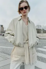 Chaqueta estilo bomber de cuero sintético en color beige, con cuello solapa, cierre frontal con cremallera, mangas largas y bajo elástico fruncido.