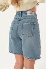 Bermuda de denim en tono azul medio, con tiro medio, cierre frontal con cremallera y botón, y diseño de cinco bolsillos.