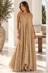 Vestido largo color dorado, confeccionado en tela semitransparente con diseño bordado floral y detalles de brillo. Presenta escote en V, tirantes finos ajustables con borlas en los hombros, y corte amplio y fluido con gran movimiento.
