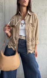 Sobrecamisa de gamuza color beige, con cuello camisero, cierre frontal con botones metálicos y bolsillo en el pecho. Presenta un diseño con calados florales y bordes ondulados en la parte inferior.