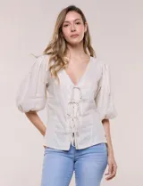 Blusa color beige confeccionada en lino, con escote en V pronunciado y cierre frontal de múltiples lazos para atar. Presenta mangas abullonadas tres cuartos con puños elásticos y corte relajado.