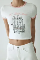 Remera corta entallada de algodón, color blanco, con ribetes rosados en el cuello y mangas, y estampa frontal de un edificio parisino con la inscripción "L'Hôtel Fleuri".