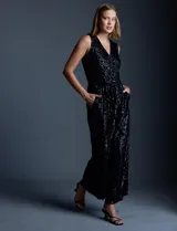 Enterito de lentejuelas marca Ninety, color negro, con escote en V cruzado y diseño sin mangas. Presenta cintura elastizada con lazo de ajuste, bolsillos frontales y abertura en la espalda con cierre invisible. Incluye forro interno a tono.