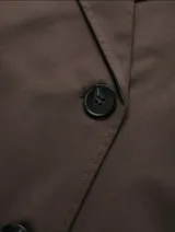 Chaqueta corta de color marrón chocolate, con diseño de doble botonadura, solapas amplias y charreteras en los hombros. Presenta mangas largas con puños ajustables mediante trabillas y bolsillos laterales integrados.