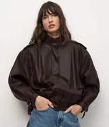 Campera de estilo bomber oversized confeccionada en poliuretano color marrón oscuro. Presenta cuello alto, cierre frontal, pliegues decorativos en el frente y la espalda, y puños ajustables con hebillas metálicas.