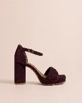 Sandalia de tacón alto con plataforma, confeccionada en gamuza color vino. Presenta un diseño de tira frontal torcida, talón cubierto y cierre con pulsera ajustable al tobillo mediante hebilla metálica. Cuenta con un taco cilíndrico robusto.