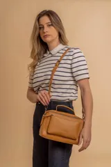 Bandolera de diseño rectangular en color suela, confeccionada en material sintético. Cuenta con un asa superior fija, correa larga ajustable y extraíble, y dos bolsillos frontales con cierre metálico, uno de ellos con diseño superpuesto. Presenta el logo de la marca grabado en el frente.