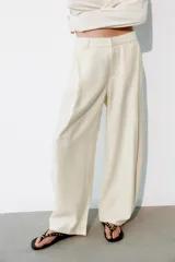 Pantalón fluido de color blanco, de tiro alto con trabillas y pliegues en el delantero. Cuenta con bolsillos laterales, cierre frontal con cremallera, botón interior y gancho metálico.