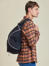 Mochila tipo bandolera (sling bag) de color negro con detalles de ribetes blancos. Presenta un diseño ergonómico de una sola correa, bolsillo frontal con cierre y logo circular de la marca Rusty. Incluye hebilla de ajuste lateral.