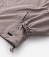 Campera rompevientos de color gris oscuro, con capucha ajustable mediante cordones, cierre frontal por cremallera y bolsillos laterales con cierre. Presenta puños elásticos y un diseño liso y minimalista.