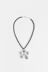 Collar con cordón trenzado negro y dije colgante en forma de flor plateada con textura martillada. Cuenta con cierre de mosquetón y cadena de alargue metálica.