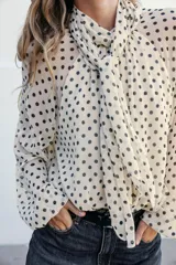 Blusa de manga larga, color marrón oscuro con estampado de lunares blancos. Presenta cuello alto con lazo ajustable en la espalda y puños elásticos.