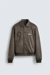 Cazadora estilo varsity confeccionada en tejido efecto ante color marrón. Presenta cuello solapa, cierre frontal con botones a presión, bolsillos laterales y puños con acabado en rib. Incluye parches bordados decorativos en el pecho.