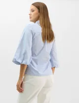Camisa de algodón con patrón de rayas verticales, cuello camisero clásico y cierre frontal con botones. Presenta detalles metálicos aplicados en el frente y mangas, con mangas 3/4 abullonadas y ruedo recto.