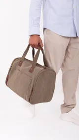 Bolso de lona color verde oliva con detalles en cuero marrón. Tiene apertura superior con cierre, bolsillos tipo guantera al frente, bolsillos interiores y amplios bolsillos laterales con cierre. Incluye doble correa de mano con agarradera forrada y correa larga desmontable y regulable.