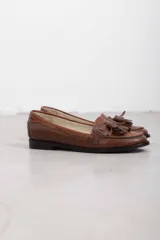 Mocasines de cuero marrón con diseño clásico, detalles de flecos y borlas en el empeine, y suela plana.
