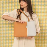 Cartera tipo tote de cuero sintético con diseño combinado en tono gris y blanco, textura granulada, asas largas para el hombro y cierre superior.