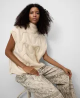 Chaleco de punto color crema, con cuello chimenea y diseño de ochos trenzados. Presenta mangas sisa con hombro caído.