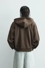 Sudadera con capucha y cierre de cremallera frontal, color marrón oscuro, de corte holgado (boxy fit) y manga larga. Presenta bolsillos frontales de vivo y acabados en rib en puños y bajo.