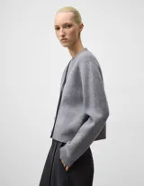 Cárdigan corto de lana cocida en color gris melange, con cuello en V, manga larga y cierre frontal mediante botones. Presenta terminaciones en punto canalé en puños y bajo.