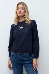 Blusa blanca de manga larga con cuello redondo, confeccionada en tela liviana con detalles de encaje vertical y bordado floral en el pecho.