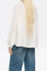 Camiseta de manga larga color blanco roto, con cuello bote y corte holgado. Presenta pespuntes en color terracota visibles en el ruedo y puños. Pequeño bordado de la marca en la parte inferior derecha.