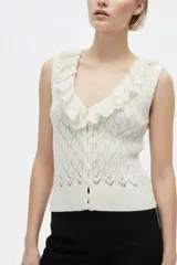 Top de punto color blanco natural, sin mangas, con escote en V pronunciado decorado con volados fruncidos. Presenta cierre frontal con pequeños botones y diseño texturizado en rombos.