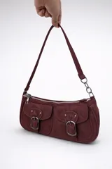 Cartera de hombro tipo baguette color bordó, confeccionada en material con textura gamuzada. Presenta dos bolsillos frontales con solapa y cierre de hebilla metálica, además de un compartimento principal con cierre. Incluye una correa fija con detalle de cadena metálica en uno de sus extremos.