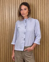 Camisa de denim y algodón a rayas celestes y blancas, con cuello alto con volados, mangas tres cuartos con puños fruncidos y cierre frontal de botones oscuros.