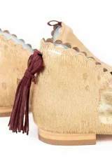 Botín de piel con acabado de pelo, color beige con estampado de manchas doradas. Presenta un diseño con bordes troquelados, aberturas laterales y detalle de borlón trasero en color granate. Incluye cuña interna y suela de cuero.