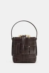 Bolso tipo bucket de diseño trenzado en color marrón oscuro con costuras a la vista en contraste. Cuenta con asa de mano fija, correa bandolera extraíble y compartimento interior de tela en color crudo con cierre mediante fruncido.