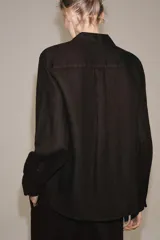 Camisa de lino color negro, de manga larga, con cuello solapa y cierre frontal con botones. Presenta un bolsillo de parche en el delantero.