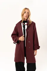 Trench de corte holgado color bordó, con cuello camisero, cierre frontal con botones y cinturón ajustable con hebilla. Presenta mangas largas con detalles de correas en los puños y bolsillos laterales.