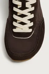 Zapatilla deportiva estilo barefoot, color marrón, con diseño de paneles en combinación de materiales y detalles en serraje. Cuenta con puntera redondeada y ancha, cordones en color crema y suela fina de goma flexible en contraste.