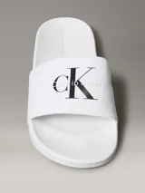 Ojotas tipo slide Calvin Klein Jeans, color blanco, con banda superior de lona de algodón que presenta el monograma de la marca en color negro. Posee suela de EVA con plantilla ergonómica moldeada.