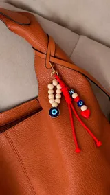 Cartera tipo hobo de cuero sintético color suela, con interior espacioso y costuras decorativas frontales. Incluye asa de hombro fija, monedero rectangular colgante a juego y un llavero amuleto con cuentas de madera, ojos turcos y detalles en rojo.