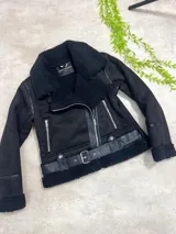 Campera tipo gamulán de color negro, confeccionada en gamuza sintética con interior y cuello de corderito. Presenta cierre frontal cruzado, bolsillos laterales con cierre, detalles en cuero sintético y cinturón ajustable en la cintura con hebilla metálica.