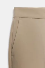 Pantalón de tiro alto color beige, de corte recto y largo tobillero, con bolsillos laterales y cierre frontal con cremallera y ganchos metálicos.