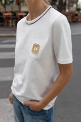 Camiseta negra de algodón, corte regular, con cuello redondo ribeteado en blanco y un bolsillo frontal con un broche metálico decorativo dorado.