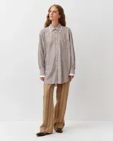 Camisa de corte recto y silueta fluida, con estampado a rayas marrones y blancas. Presenta cuello clásico, cierre frontal con botones y un bolsillo aplicado en el pecho. Las mangas largas son ligeramente abullonadas, con puños ajustados que aportan volumen a la prenda.