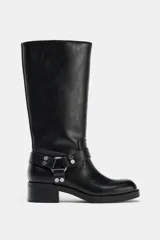 Bota estilo biker de caña media, confeccionada en cuero sintético negro. Presenta un diseño con tacón en bloque, punta cuadrada y detalle de doble tira con tachas metálicas y arandela en la zona del tobillo.