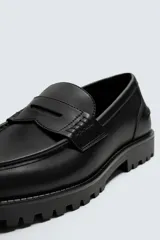 Mocasín de vestir negro, estilo penny loafer con antifaz en el empeine. Presenta forro y plantilla interior a tono, y suela track gruesa y dentada.
