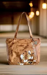 Cartera tipo shopper de cuero suave color marrón terra, con diseño pintado a mano que incluye salpicaduras y la inscripción de la marca. Cuenta con dos asas largas para llevar al hombro, cierre principal, forro interior con bolsillo y un dije metálico colgante.