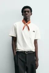 Camiseta de corte relajado (relaxed fit) color blanco hueso, con cuello redondo, manga corta y bolsillo plastrón en el pecho. Presenta un bordado pequeño y contrastante de una palmera en el área del bolsillo.