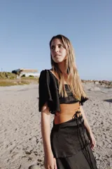 Vestido largo negro de dos piezas, compuesto por un top con escote amplio y mangas cortas amplias, y una pollera larga con tajo lateral. Incluye una faja marrón de cuero que marca la cintura.