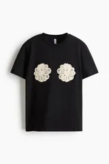 Remera negra de punto de algodón, de corte holgado con hombros caídos, cuello redondo acanalado y mangas cortas. Presenta dos apliques decorativos circulares de crochet en tono crudo a la altura del pecho.