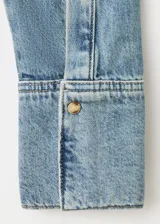 Camisa de denim estilo vaquero, con cuello camisero, manga larga con puños abotonados, cierre frontal con botones y detalle de volantes en el frente.