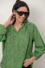 Camisa de algodón verde con bordados al tono, escote en V y cuello camisero corto, mangas tres cuartos abullonadas con puños elásticos y cierre frontal parcial con botones.